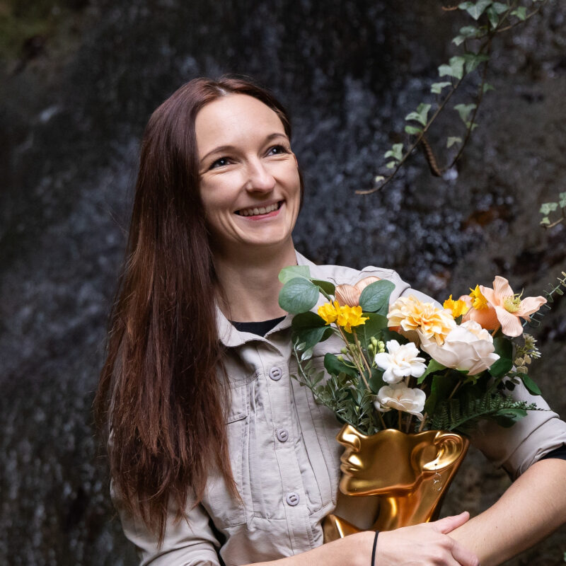 Michaela Baumgartner, Dipl. Mentaltrainerin, in der Natur, in der Hand hält sie einen goldenen Kopf wo Blumen rauswachsen, als Sinnbild für die positiven Gedanken